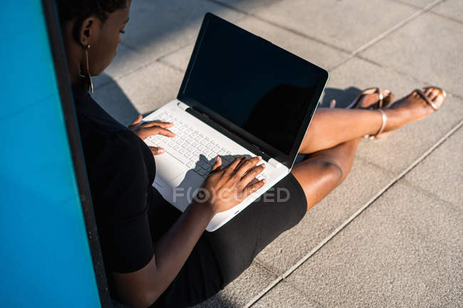 Mujer afroamericana concentrada en elegante vestido negro usando computadora portátil mientras se relaja en el pavimento en la calle - foto de stock