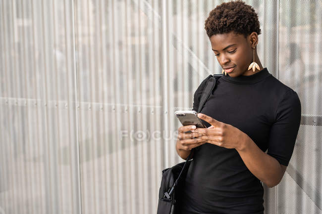 Орієнтовані стильний афроамериканець жінка в чорному плаття повідомленнями смартфон, стоячи на металевому фоні — стокове фото