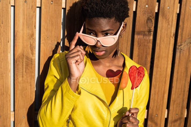 Mujer afroamericana de moda en gafas de sol en chaqueta amarilla disfrutando de piruleta en forma de corazón por valla de madera - foto de stock