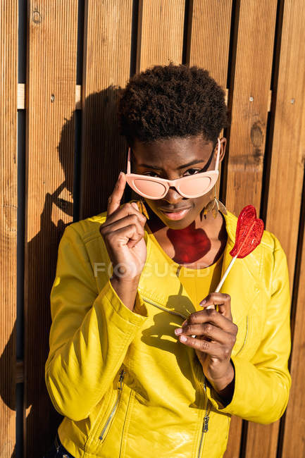 Trendy African American woman in sunglasses in yellow jacket enjoying heart shaped lollipop by wooden fence — Stock Photo