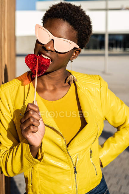 Trendy donna afroamericana in occhiali da sole in giacca gialla godendo di lecca-lecca a forma di cuore da recinzione in legno — Foto stock