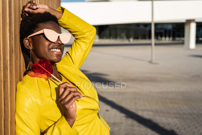 Vista laterale della donna afroamericana alla moda in occhiali da sole in giacca gialla godendo di lecca-lecca a forma di cuore appoggiata alla recinzione in legno — Foto stock