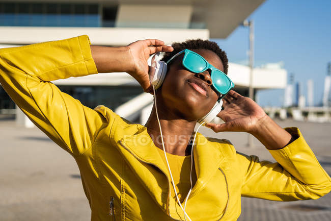 Femme afro-américaine dans une veste lumineuse élégante et des lunettes de soleil bleu vif à l'aide d'écouteurs debout près d'un bâtiment moderne — Photo de stock