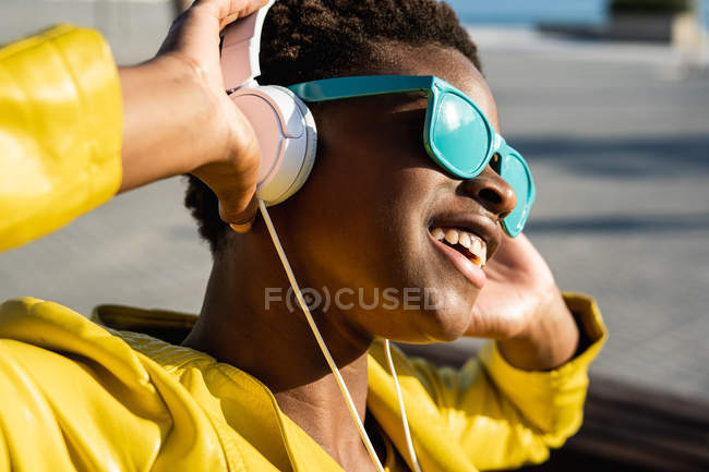 African American woman in stylish blue sunglasses using headphones standing near a modern building — Stock Photo