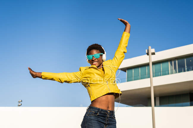 Bajo ángulo de la mujer afroamericana feliz en chaqueta brillante con estilo y gafas de sol saltando con las manos arriba - foto de stock