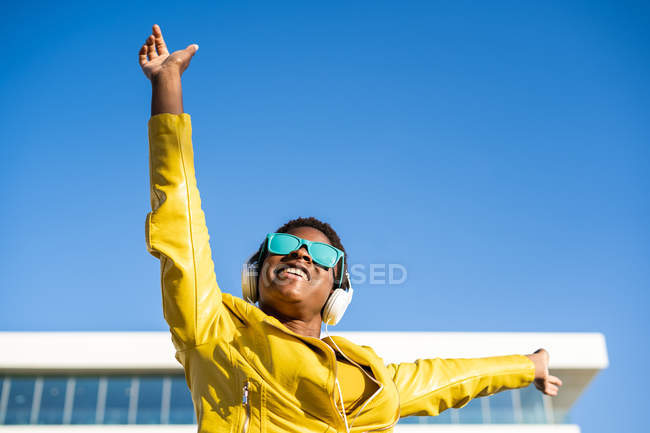 Angolo basso di felice donna afroamericana in elegante giacca luminosa e occhiali da sole saltando con le mani in alto — Foto stock