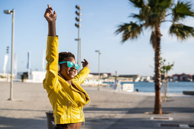 Low angle of happy African American woman in stylish bright jacket and sunglasses jumping with hands up on street — Stock Photo
