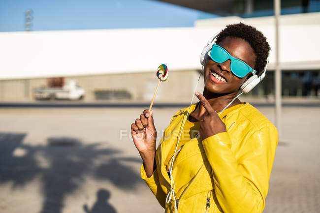 Mulher afro-americana em óculos de sol em casaco amarelo desfrutando de um pirulito e ouvindo música em fones de ouvido — Fotografia de Stock