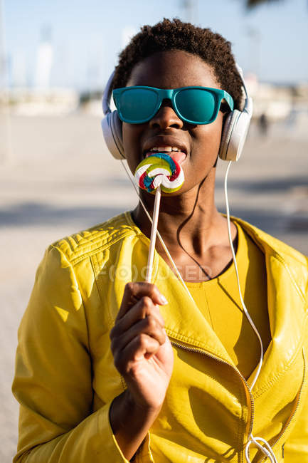 African American woman in sunglasses in yellow jacket enjoying a lollipop and listening to music on headphones — Stock Photo