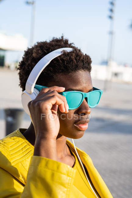 African American woman in stylish bright jacket and bright blue sunglasses using headphones standing near a modern building — Stock Photo