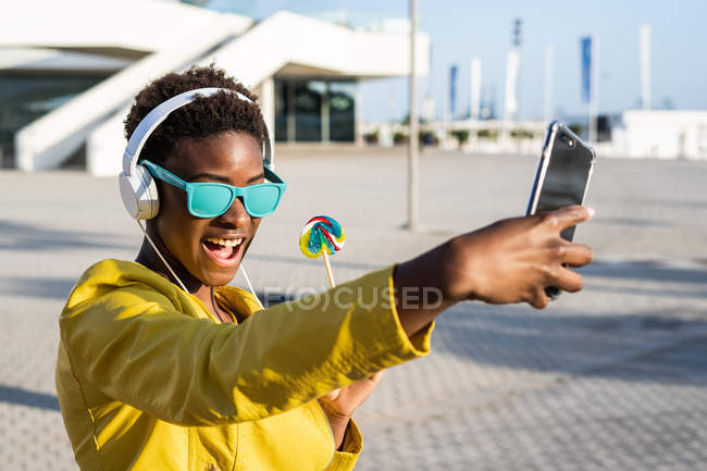 Mujer afroamericana disfrutando de la piruleta y escuchando música en los auriculares mientras se toma una selfie en un teléfono móvil - foto de stock