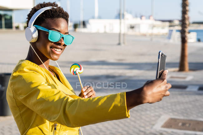 Mujer afroamericana disfrutando de la piruleta y escuchando música en los auriculares mientras se toma una selfie en un teléfono móvil - foto de stock