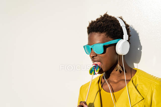 Mujer afroamericana con gafas de sol en chaqueta amarilla disfrutando de una piruleta y escuchando música en los auriculares - foto de stock