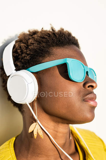 Mujer afroamericana con elegante chaqueta brillante y gafas de sol azul brillante usando auriculares apoyados en una pared blanca - foto de stock