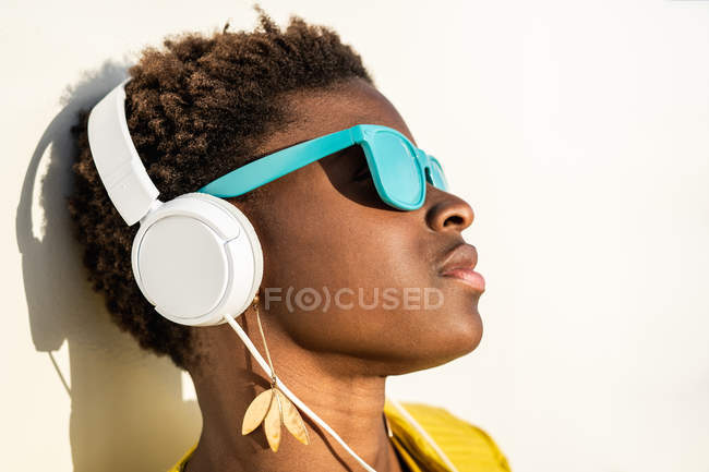 Mulher afro-americana em elegante jaqueta brilhante e óculos de sol azul brilhante usando fones de ouvido encostados em uma parede branca — Fotografia de Stock