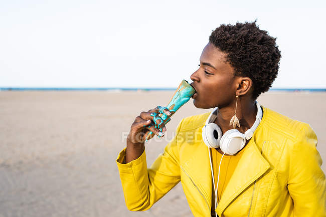 Vista laterale della donna afroamericana alla moda in giacca gialla che si gode il gelato in piedi in una spiaggia di sabbia — Foto stock