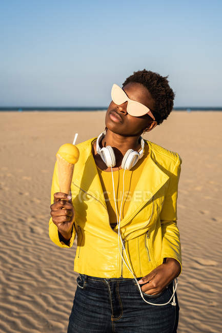 Mujer afroamericana de moda en auriculares y gafas de sol en chaqueta de color amarillo brillante disfrutando de helado en el desierto - foto de stock