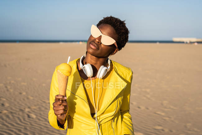 Trendy donna afroamericana in occhiali da sole in giacca gialla godendo di gelato in piedi sulla spiaggia di sabbia — Foto stock