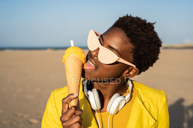 Trendy African American woman in sunglasses in bright yellow jacket enjoying ice cream standing in sandy beach — Stock Photo
