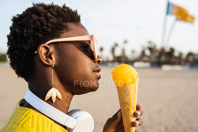 Vista lateral da mulher afro-americana na moda em casaco amarelo brilhante desfrutando de sorvete em pé na praia arenosa — Fotografia de Stock