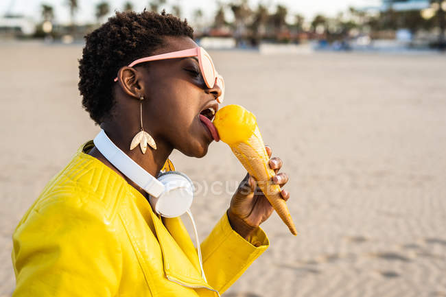 Vista lateral de la mujer afroamericana de moda en chaqueta de color amarillo brillante disfrutando de helado de pie en la playa de arena - foto de stock