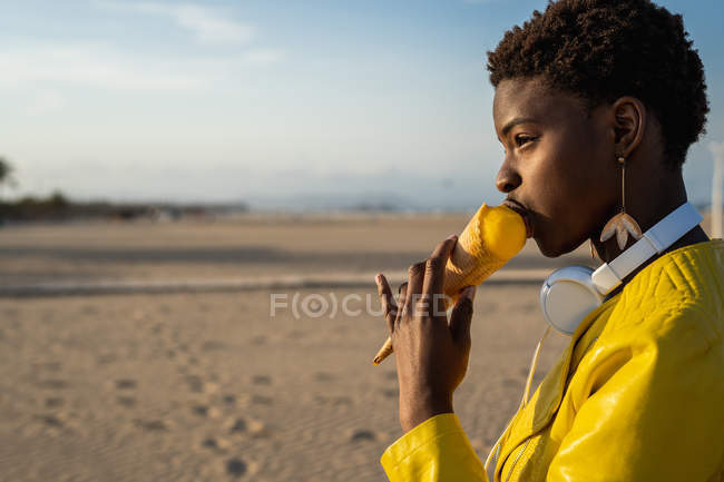 Вид сбоку на трепещущую афроамериканку в ярко-желтой куртке, наслаждающуюся мороженым, стоя на песчаном пляже — стоковое фото