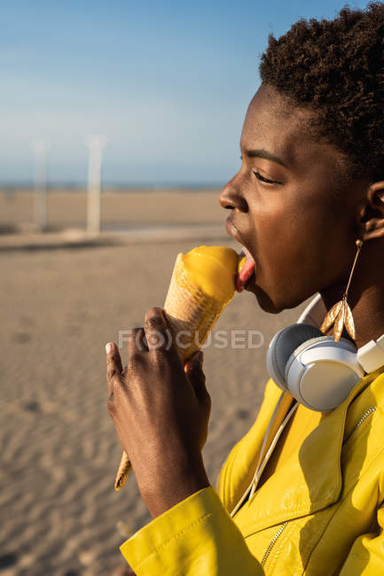 Vista lateral de la mujer afroamericana de moda en chaqueta de color amarillo brillante disfrutando de helado de pie en la playa de arena - foto de stock