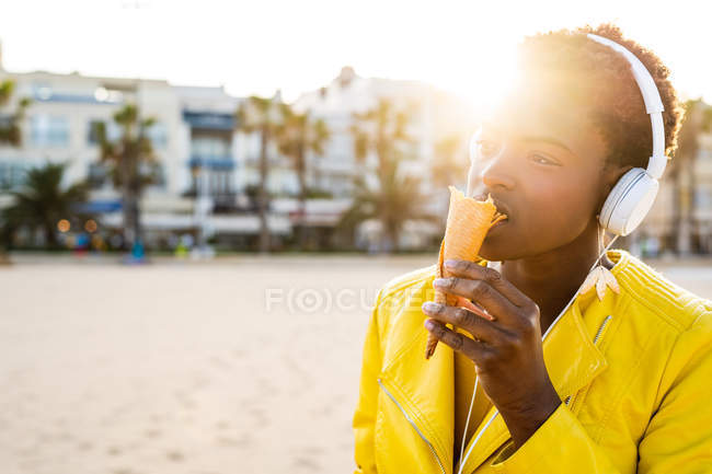 Side view of trendy African American woman in headphones in bright yellow jacket enjoying ice cream standing in sunlight — Stock Photo