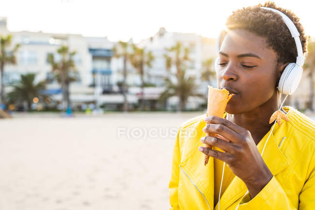 Vista lateral de la mujer afroamericana de moda disfrutando de helado con los ojos cerrados de pie en la playa de arena - foto de stock