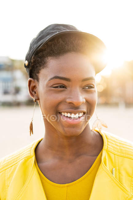 Retrato de mujer afroamericana feliz en chaqueta brillante con estilo mirando hacia otro lado en la playa de arena fondo borroso - foto de stock