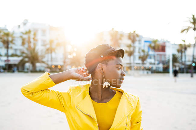 Retrato de mujer afroamericana con elegante chaqueta brillante mirando hacia otro lado en la playa de arena fondo borroso - foto de stock