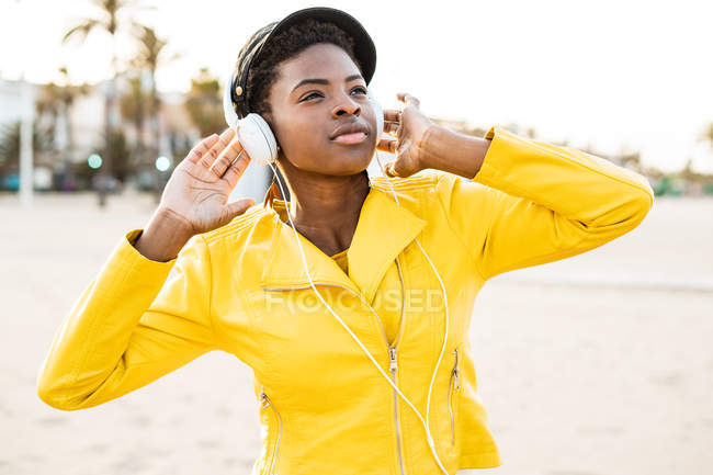 Retrato de mujer afroamericana con elegante chaqueta brillante mirando hacia otro lado en la playa de arena fondo borroso - foto de stock