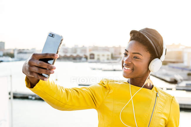 Donna afroamericana in elegante giacca luminosa scattare selfie e ascoltare musica sulle cuffie — Foto stock