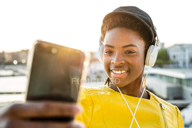 Mujer afroamericana con elegante chaqueta brillante tomando selfie y escuchando música en los auriculares - foto de stock