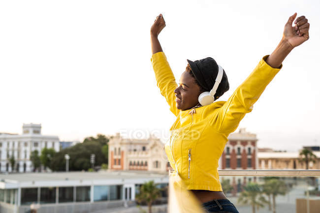 Vista lateral da mulher afro-americana na jaqueta da moda olhando para longe com os braços para cima no fundo borrado — Fotografia de Stock