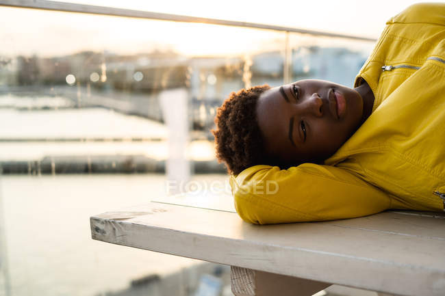 Side view of stylish black African American female in modern jacket relaxing on wooden bench and looking away — Stock Photo