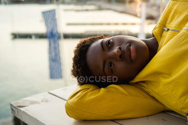 Side view of stylish black African American female in modern jacket relaxing on wooden bench and looking away — Stock Photo
