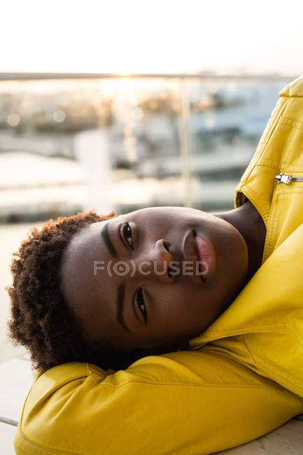 Vista lateral do elegante preto afro-americano feminino em casaco moderno relaxante no banco de madeira e olhando para longe — Fotografia de Stock