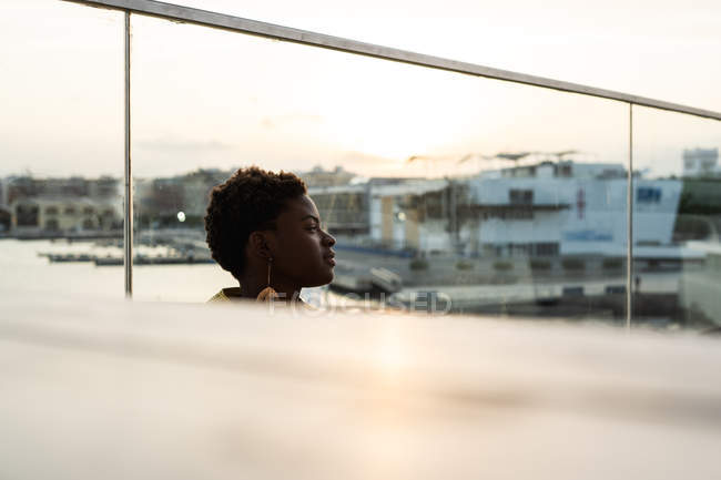 Селективный вид сбоку безмятежной афроамериканки, охлаждающейся на стеклянном балконе и отводящей взгляд — стоковое фото