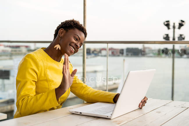 African American woman in yellow jacket using laptop web cam at wooden desk in city on blurred background — Stock Photo