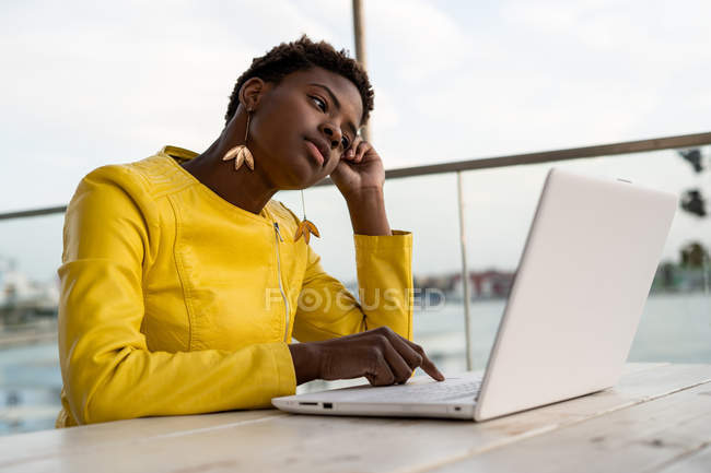 Tired African American woman in yellow jacket using laptop at wooden desk in city on blurred background — Stock Photo