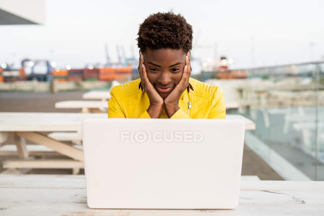 Rostro sorprendido de mujer afroamericana negra con chaqueta amarilla usando portátil en escritorio de madera en la ciudad sobre fondo borroso - foto de stock