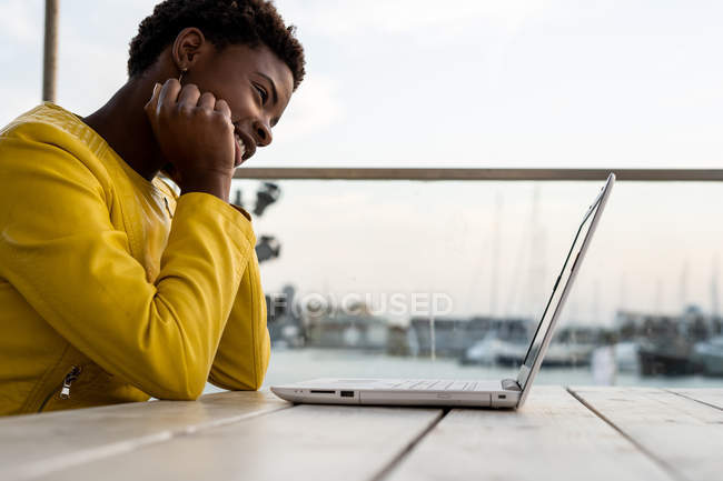 Афроамериканка в жёлтой куртке с ноутбуком за деревянным столом в городе на размытом фоне — стоковое фото