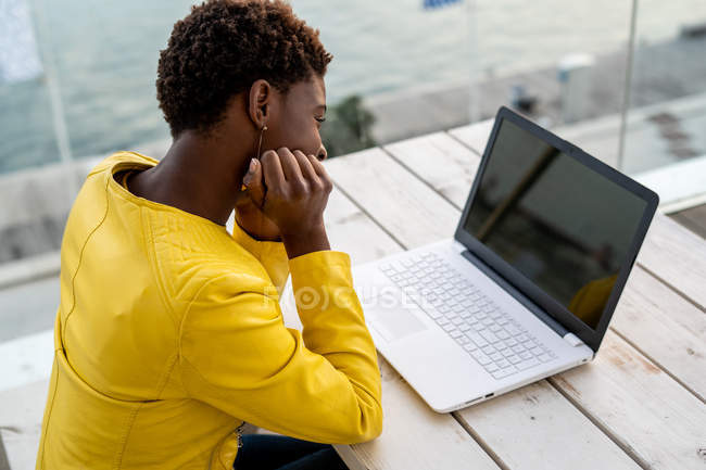 Femme afro-américaine en veste jaune à l'aide d'un ordinateur portable au bureau en bois en ville sur fond flou — Photo de stock