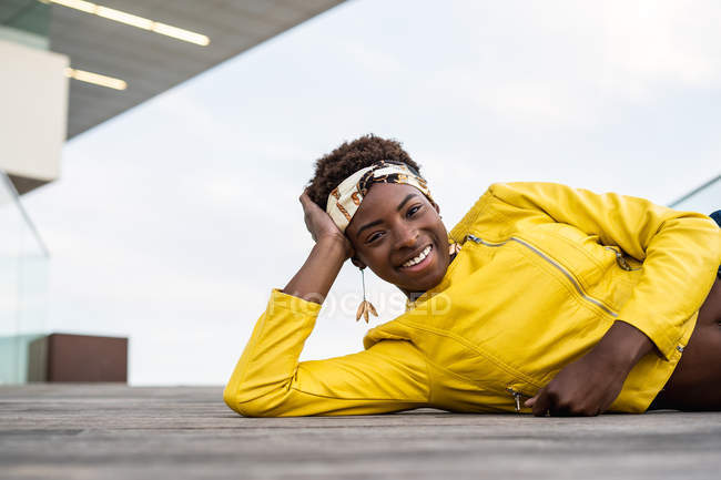 Stylische Afroamerikanerin in moderner Jacke entspannt auf Holzboden liegend und in die Kamera blickend — Stockfoto