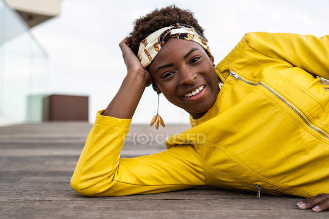 Stylish African American female in modern jacket relaxing lying down on wooden floor and looking in camera — Stock Photo