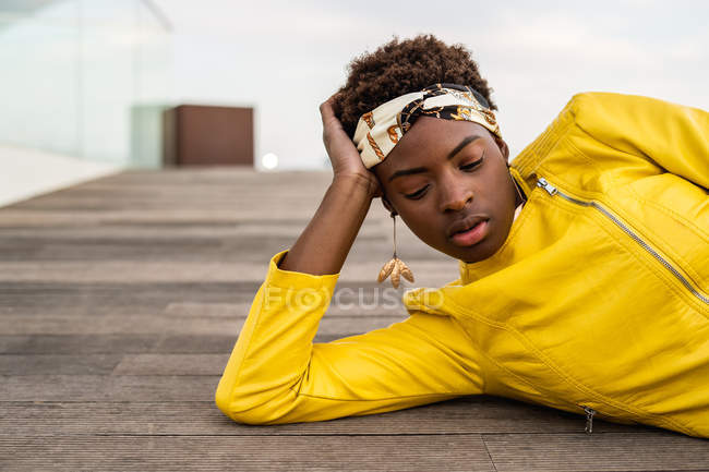 Mulher afro-americana elegante em casaco moderno relaxante deitado no chão de madeira e olhando para baixo — Fotografia de Stock
