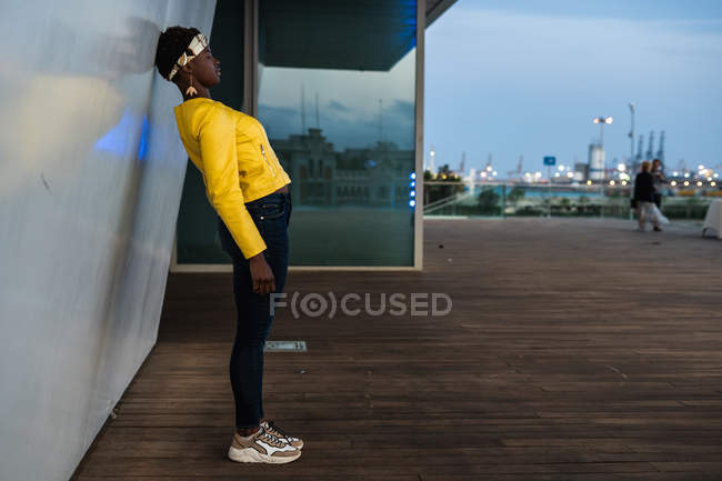 Vista lateral de la mujer afroamericana moderna apoyada en la pared del edificio moderno y soñando despierto en la ciudad al atardecer mirando hacia otro lado - foto de stock