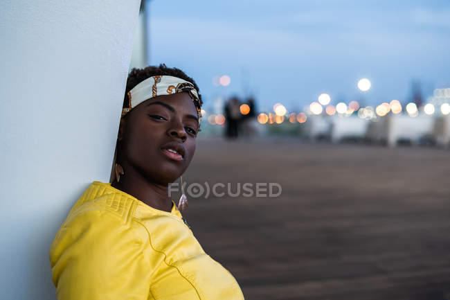 Seitenansicht einer afrikanisch-amerikanischen Frau, die sich an die Wand eines modernen Gebäudes lehnt und in die Kamera blickt — Stockfoto