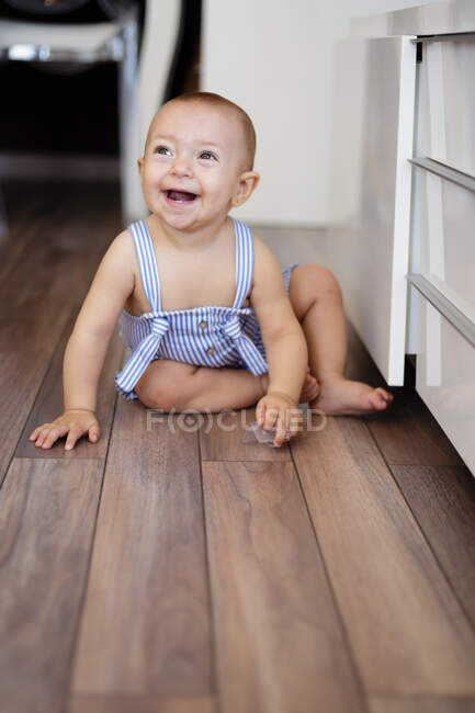 Emocionado bebé descalzo mirando hacia otro lado mientras se sienta en parquet cerca de mostradores en la acogedora cocina en casa - foto de stock